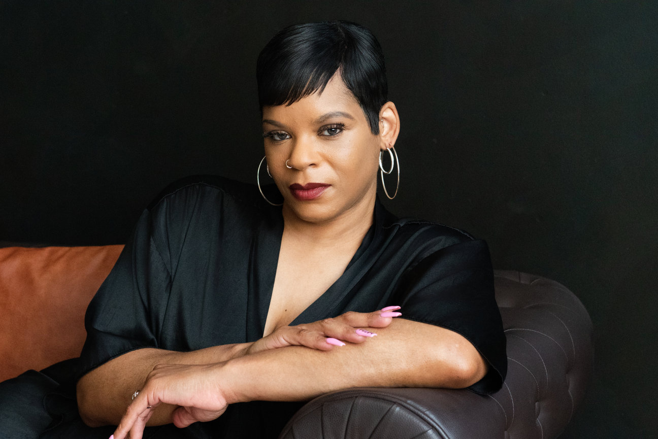 A woman with short black hair and hoop earrings sits on a leather couch, looking confidently at the camera.