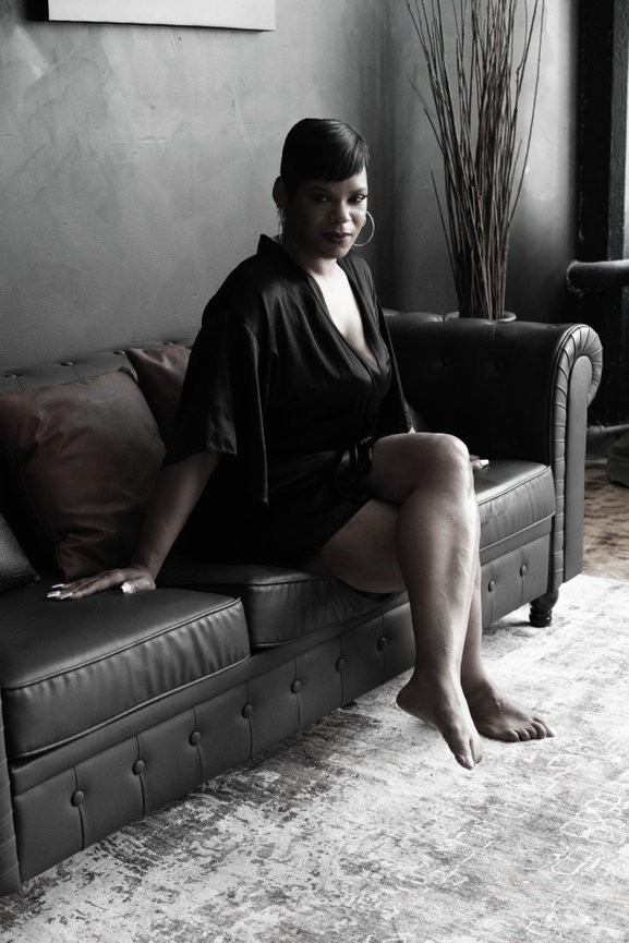 Woman in a black robe sitting on a leather sofa with a relaxed pose in a softly lit room.