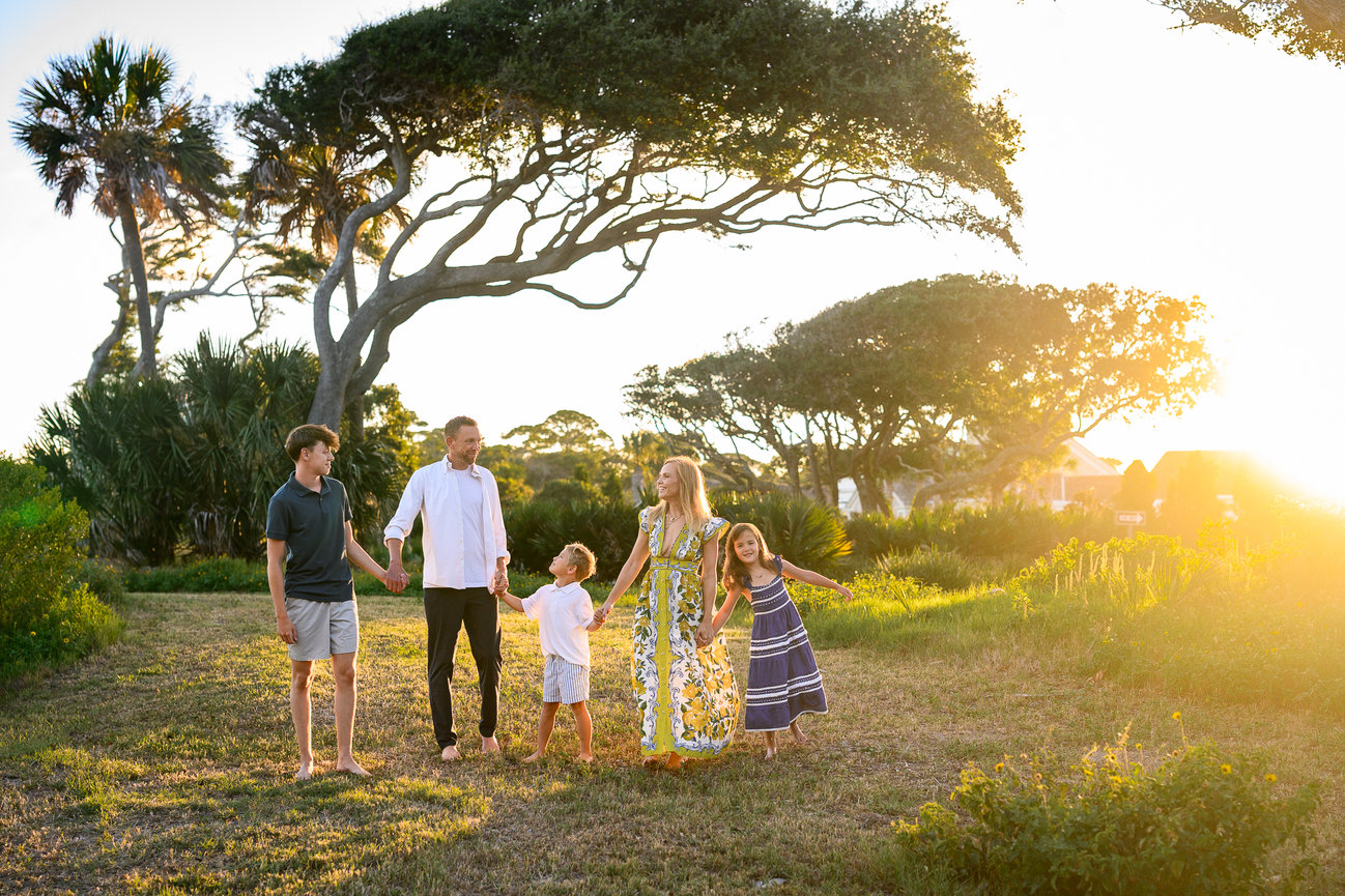 Family of five walking hand-in-hand through a sunlit, grassy field with trees in the background.