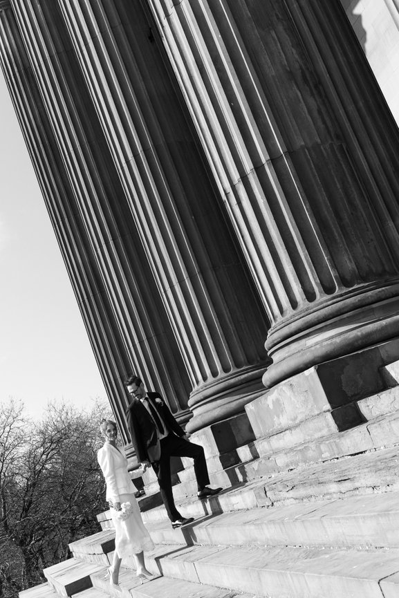Cinematic black and white wedding photography of a bride and groom walking up the grand steps of St George's Hall, Liverpool, framed by massive neoclassical columns.