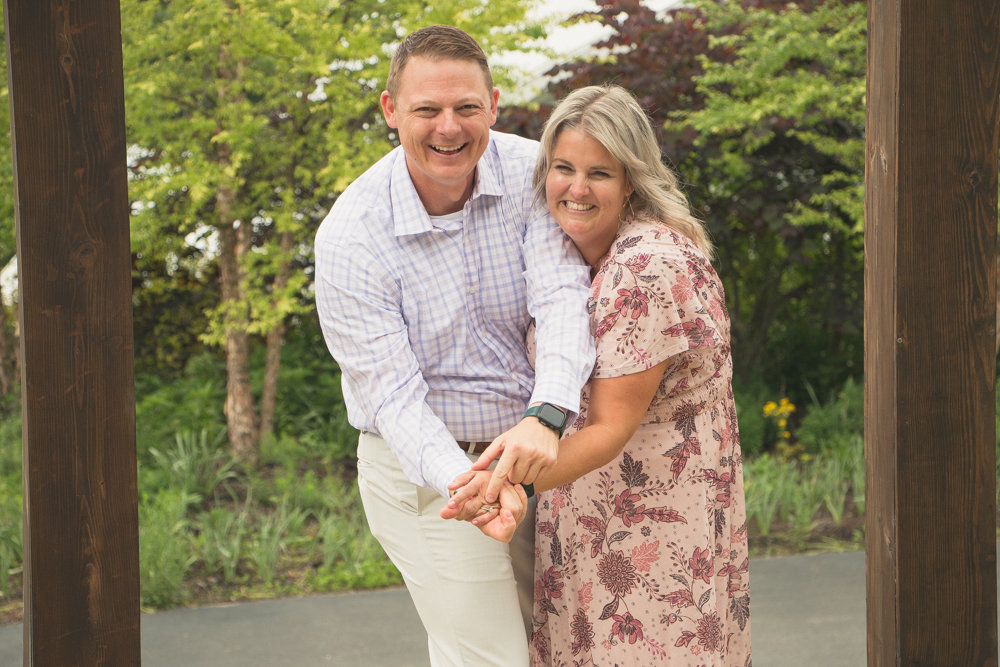 A smiling couple holding hands, standing outdoors with greenery in the background.