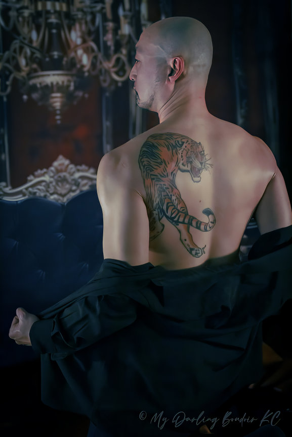 Person with a tiger tattoo on their back, holding a black jacket, standing in front of a decorative chandelier backdrop.