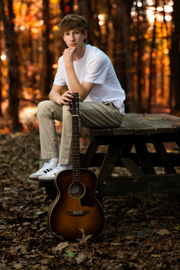 High School Senior sitting on a picnic table in a forest with an acoustic guitar in autumn light photographed by Darren Elias Photography.