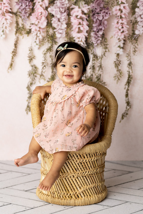 Smiling baby girl in a floral dress sitting in a wicker chair, surrounded by pastel floral backdrop.