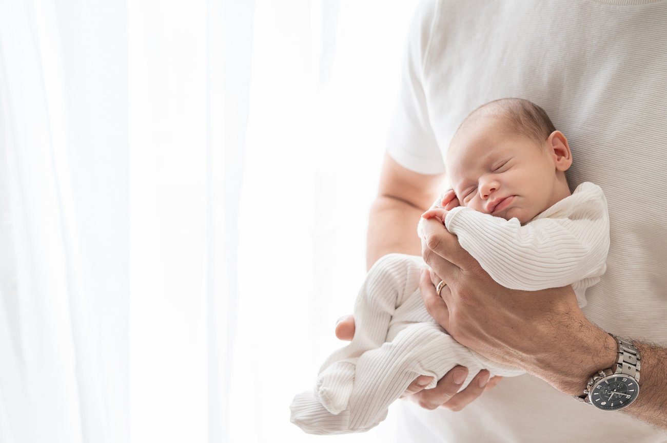 Father cradling newborn against white curtain