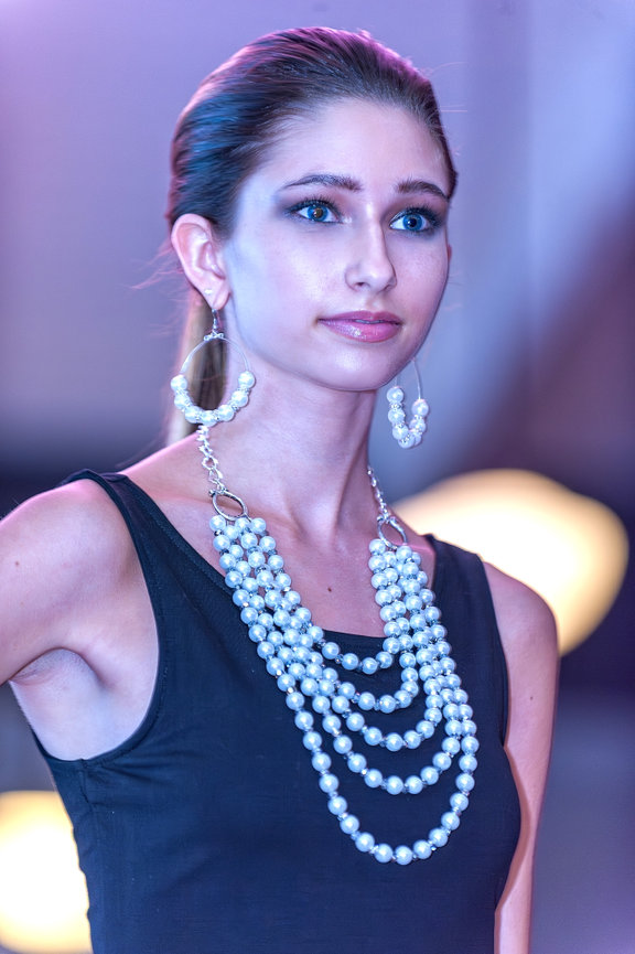Fashion model on runway wearing a black sleeveless top, oversized hoop earrings adorned with pearls, and a multi-strand pearl statement necklace, under cool-toned lighting.
