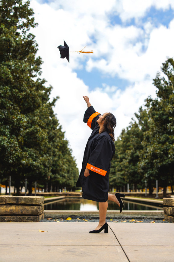 Graduate throwing cap in the air, celebrating outdoors with trees and a reflective pond in the background the University of Texas in Dallas