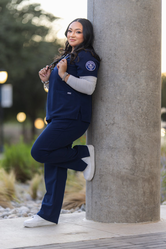Smiling woman in navy scrubs and white sneakers leaning against a column outdoors at the university of Texas at Arlington