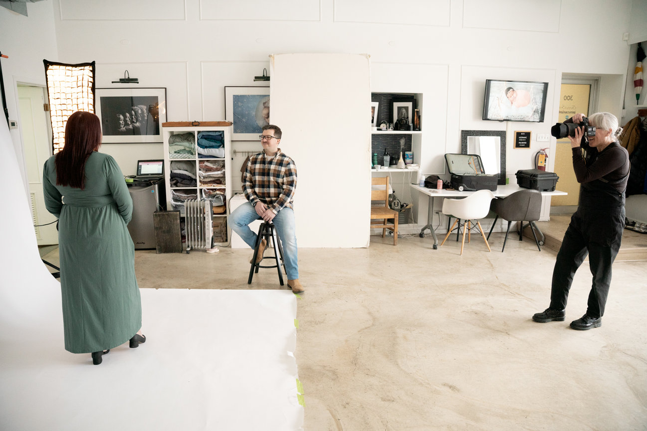 Photographer taking a picture of a seated person in a well-lit studio with various equipment and decor.