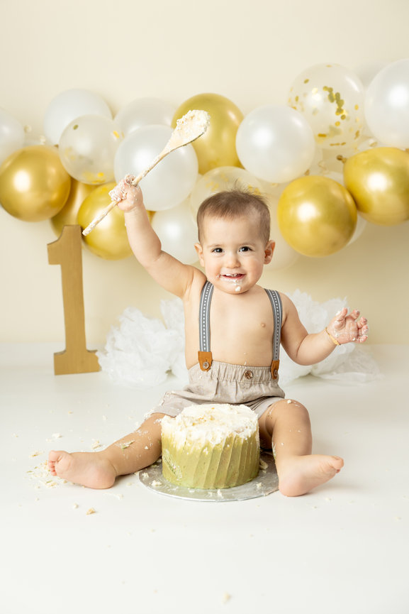 Baby celebrating first birthday with a cake smash; wearing suspenders, surrounded by gold and white balloons, number one decoration.