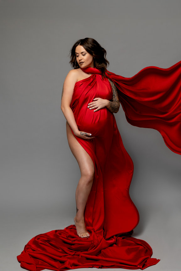 Pregnant woman in a flowing red gown poses barefoot against a gray background.