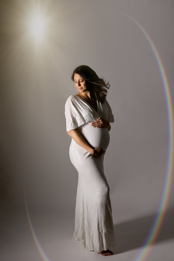 Pregnant woman in a white dress stands serenely under a soft light, with a subtle rainbow lens flare effect.
