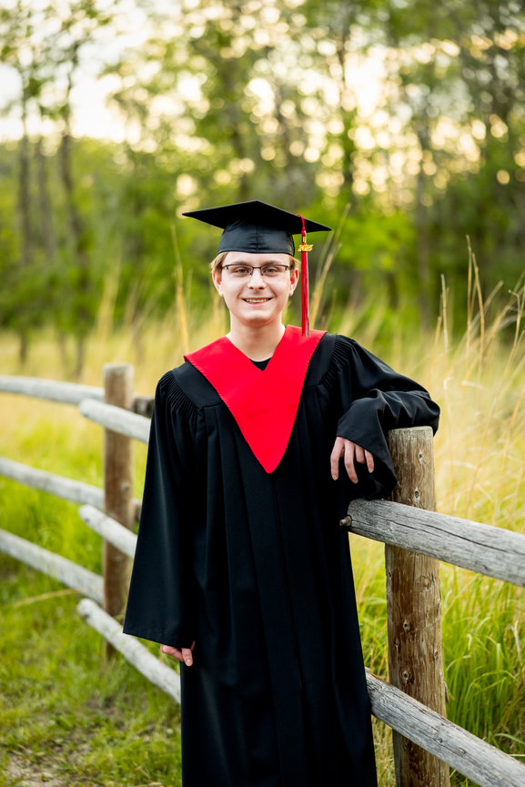 Graduate in cap and gown with red sash smiles, standing by a wooden fence in a natural setting.