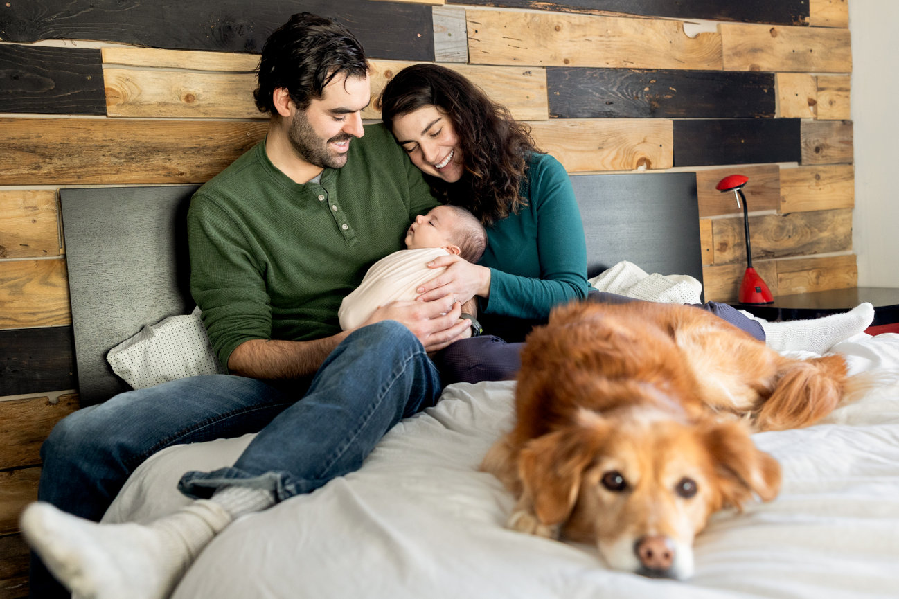 A couple sits on a bed holding a baby, with a dog lying in the foreground.