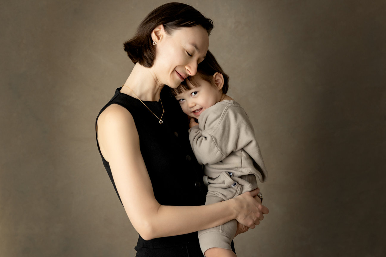 A woman lovingly holds a smiling child against a neutral background.