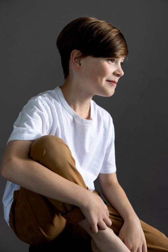 Young boy in a white shirt and brown pants sitting in profile against a gray background
