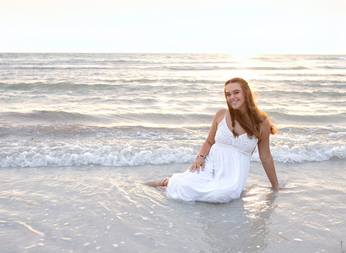 High school senior sitting in the sand during golden hour at her senior adventure destination photoshoot with Angie Vincent of Heart to Art Photography.
