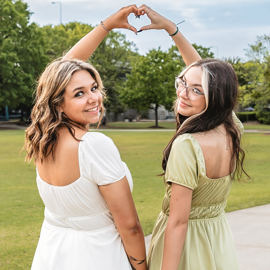 Two high school teenage girls in dresses smiling outdoors, making a heart shape with their raised hands.