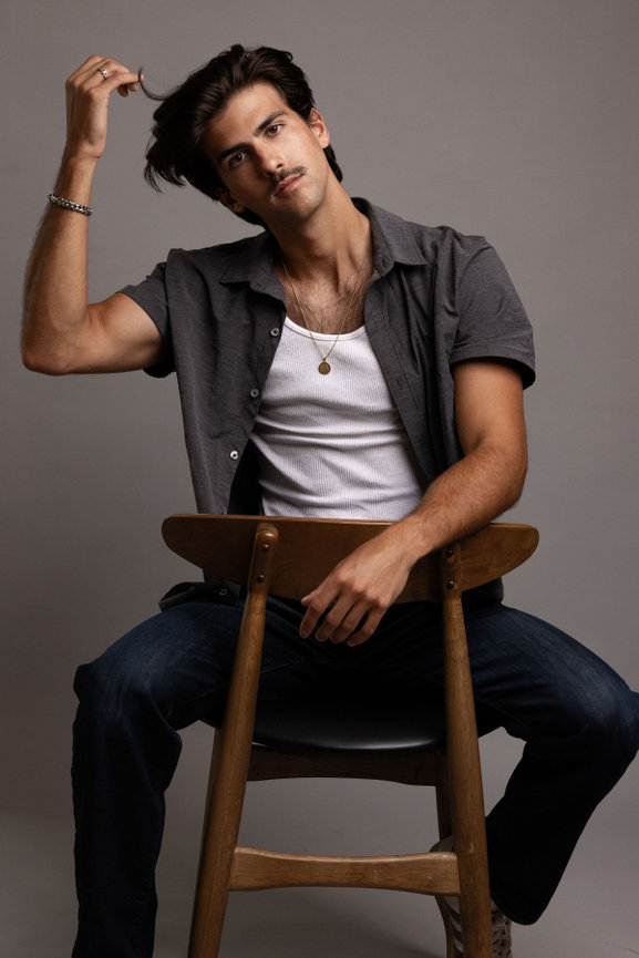 Young Male model posing on a wooden chair in a studio, wearing a gray shirt, white tank top, and jeans as he brushes his hand through his hair