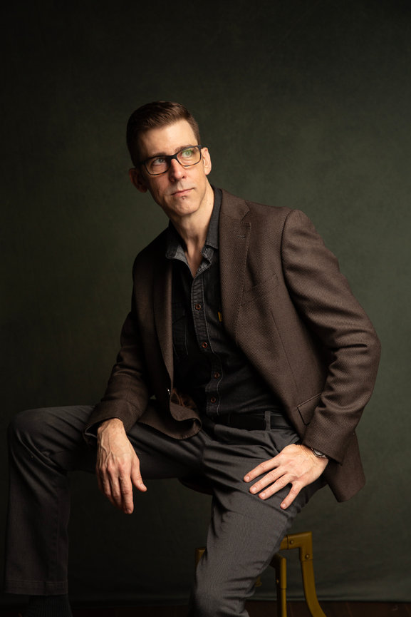 Dallas professional headshot of a man in glasses and a brown suit jacket, seated against a dark studio background.