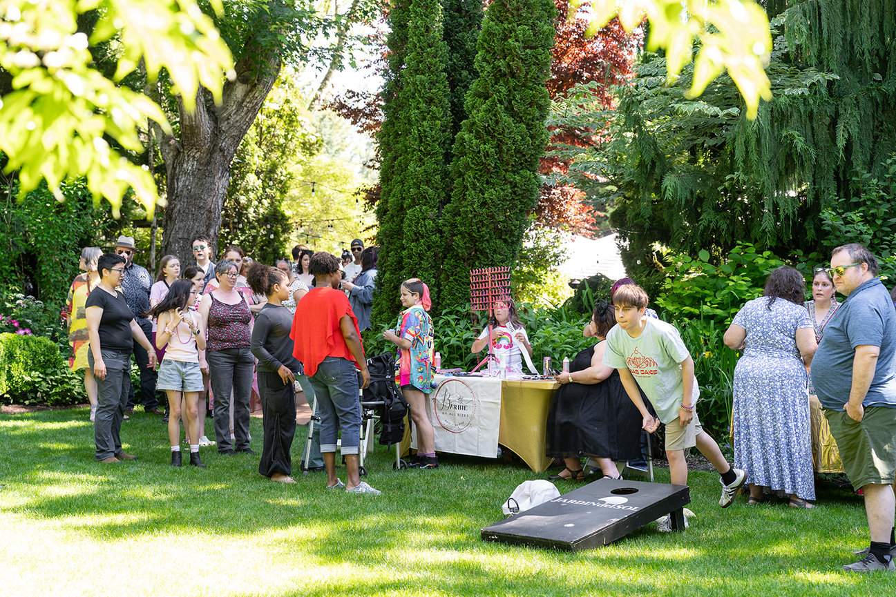 People socializing at an outdoor garden gathering with a cornhole game and vendor table.
