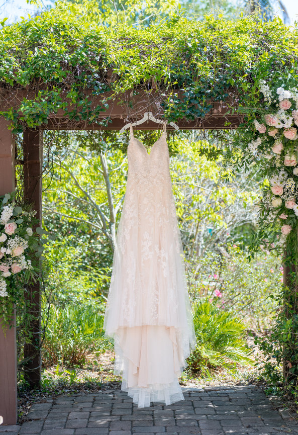 White wedding dress hanging from a floral arch in a garden.
