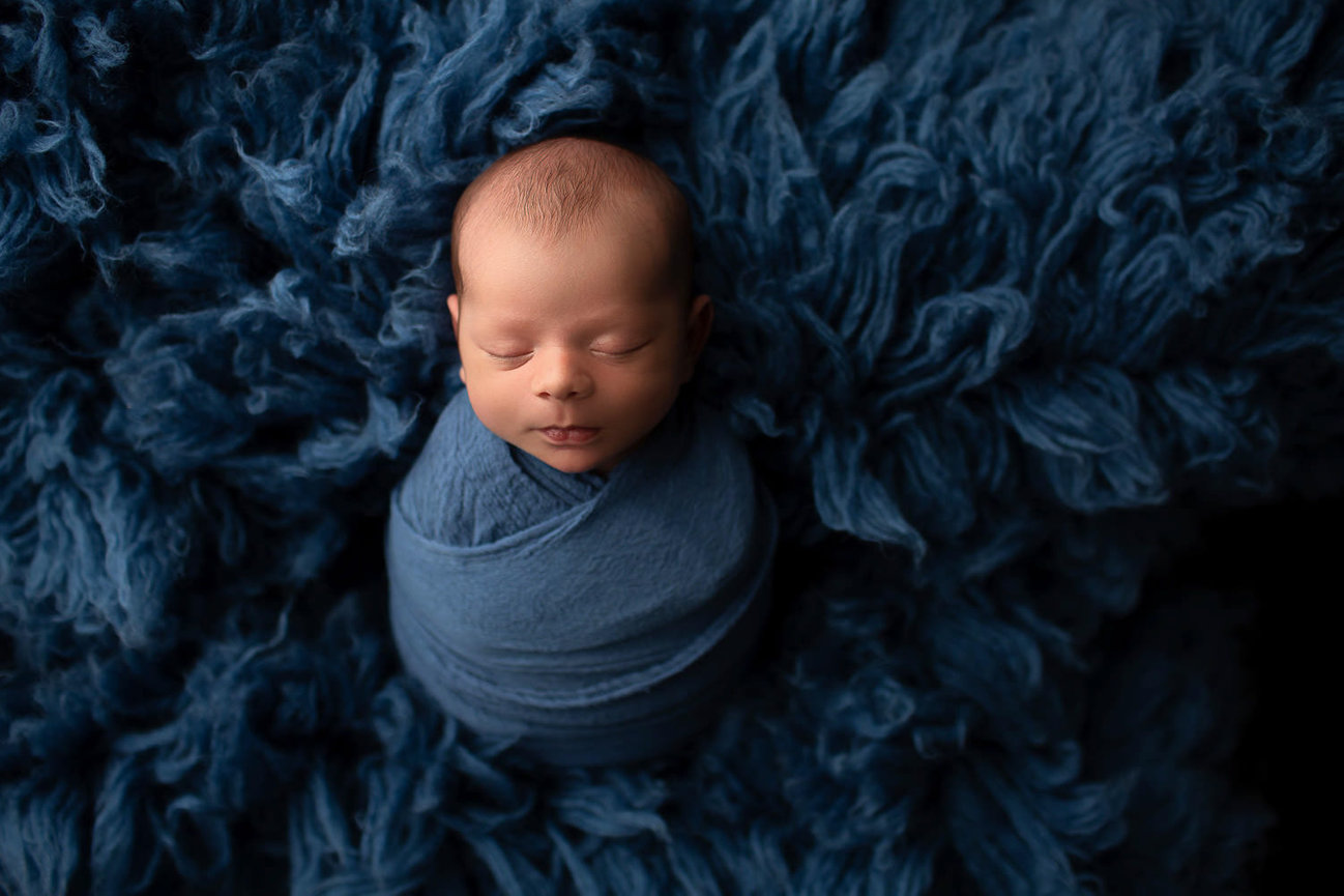 Newborn baby boy in blue wrap sleeping in potato pose on matching blue flokati during newborn photoshoot with newborn photographer in Kansas City Dreamworks Photography 