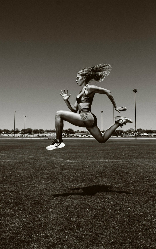 Female Athlete jumping and looking like she is running in mid air with a sport field in backdrop in photo by Sara Racey Photographer.