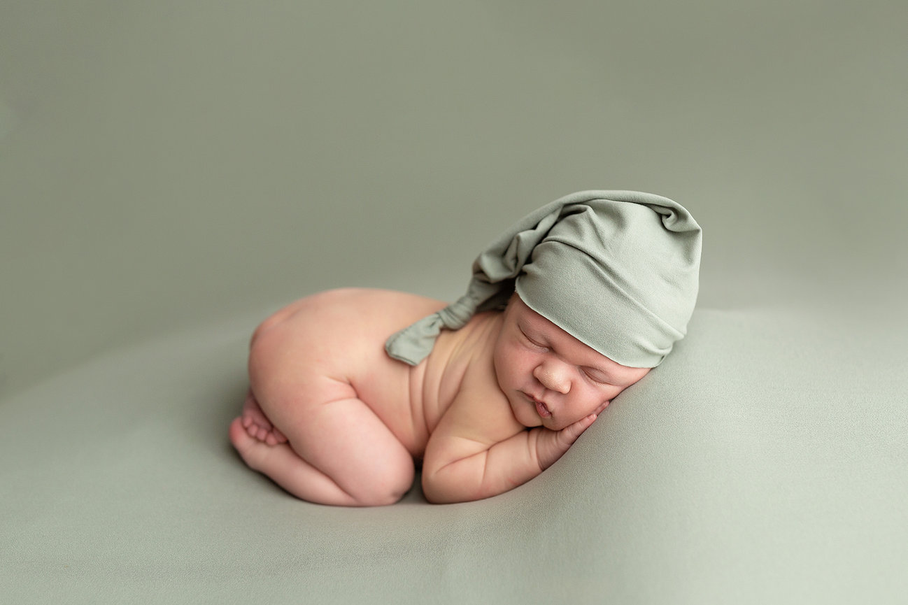 Omaha, Nebraska Newborn Photographer photographs a sleeping baby on a soft surface, wearing a light green hat.