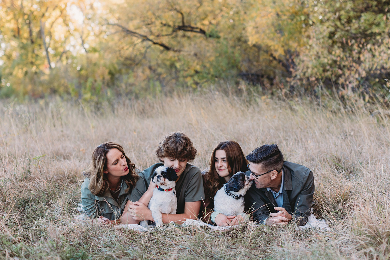 Family lying in a grassy field with two dogs, surrounded by autumn trees.