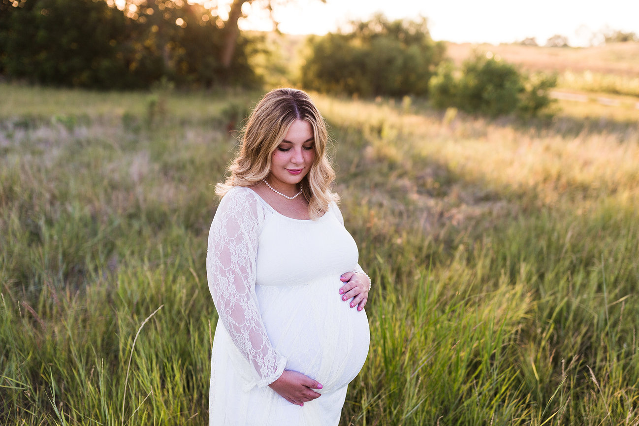 Pregnant woman in a white lace dress standing in a grassy field at sunset.