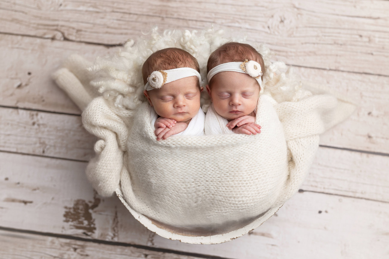 Two sleeping newborn twins wrapped in a white blanket, lying on a textured wooden surface.