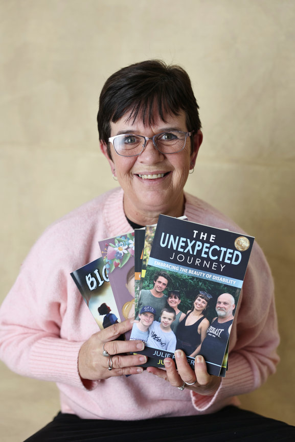 Smiling woman in glasses holding several books, including The Unexpected Journey, against a beige background. Business content photos melb