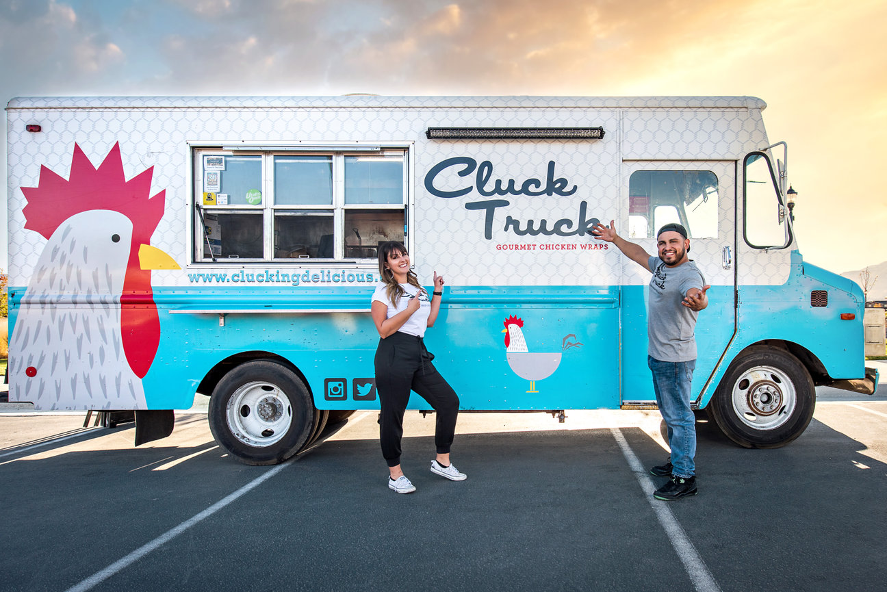 A man and woman owners of a food truck smiling and pointing to their truck.