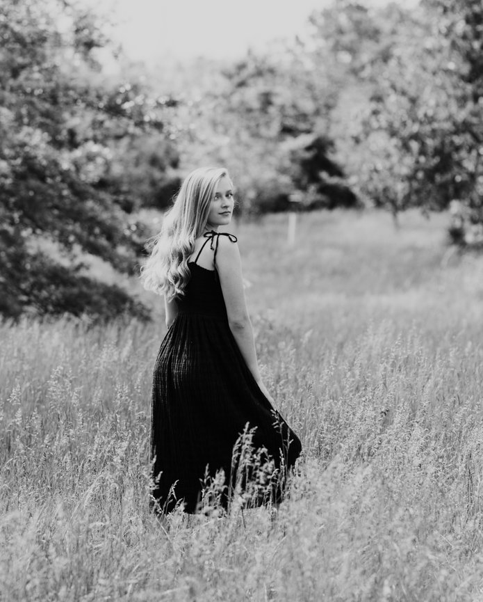A high school senior in a long dress stands in a grassy field, looking back over her shoulder. Trees and tall grass surround her. Black and white image.