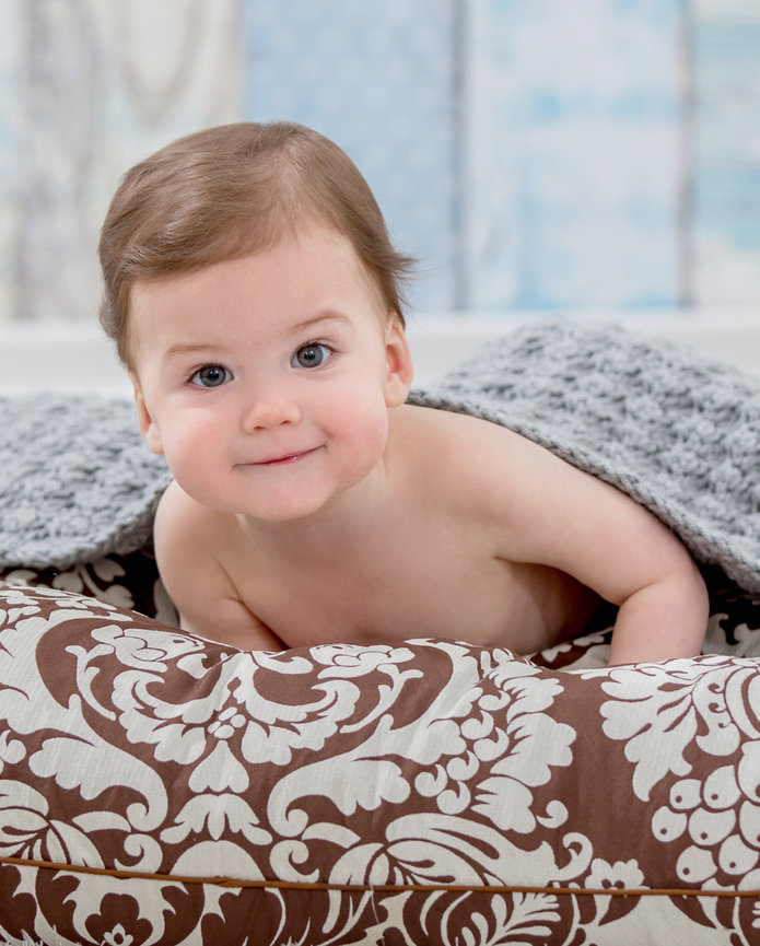 A baby smiling while lying on a patterned pillow, partially covered with a gray blanket.