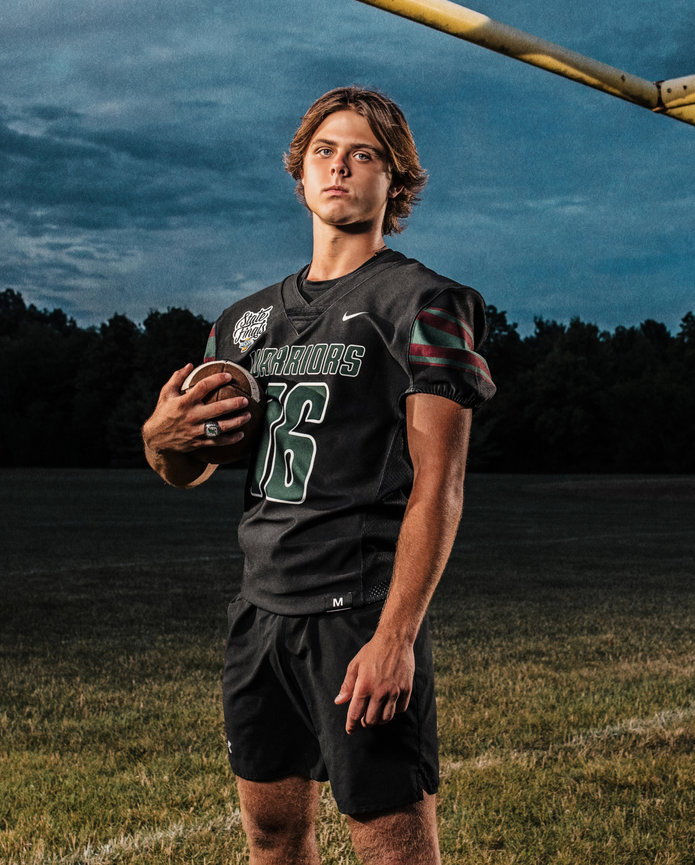 High school senior football player stands on a field under a goalpost, wearing number 6. The cloudy sky looms in the background.