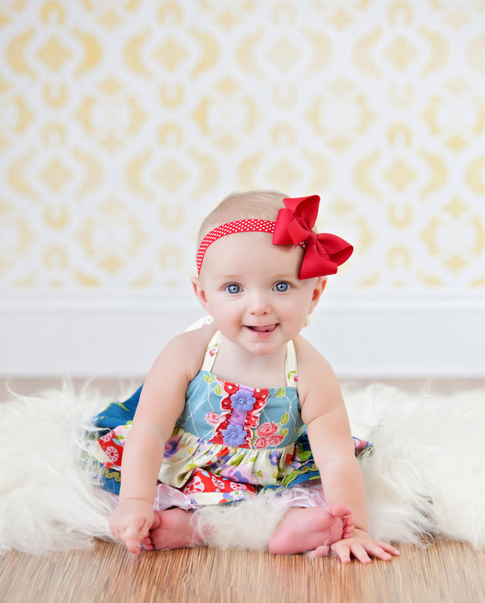 Baby in a colorful dress and red bow headband, sitting on a fluffy rug with a yellow patterned wallpaper background.