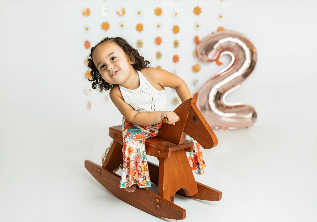Toddler sitting on wooden rocking horse in natural light studio with 2 balloon