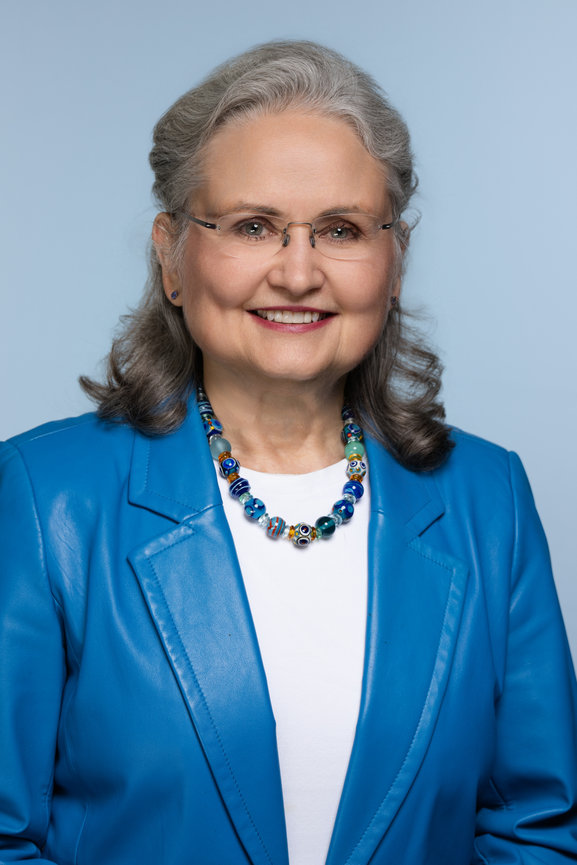 Woman in a blue blazer and necklace, smiling against a light blue background.