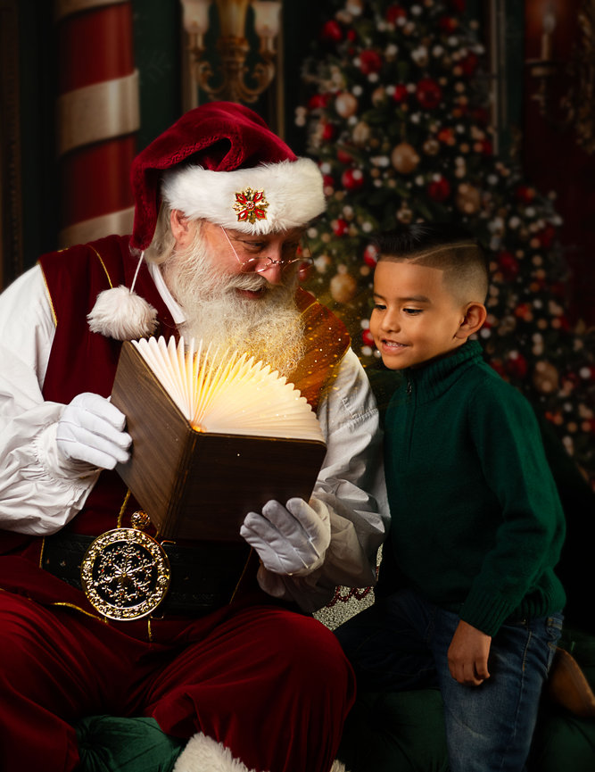 Santa reads a glowing magical book to a young boy beside a decorated Christmas tree during an Annapolis Christmas mini session.