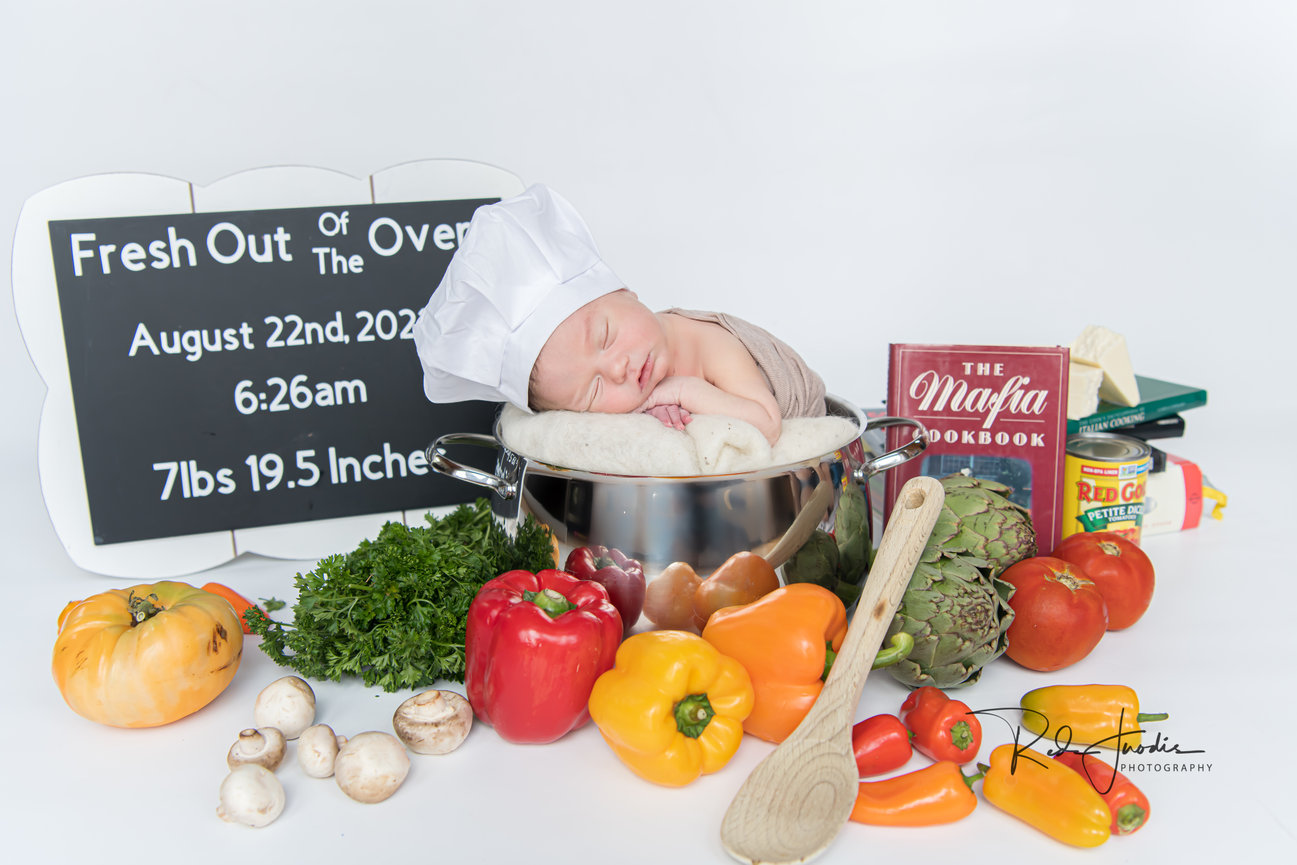 Newborn baby in a chef hat nestled in a pot, surrounded by colorful vegetables and a cookbook.