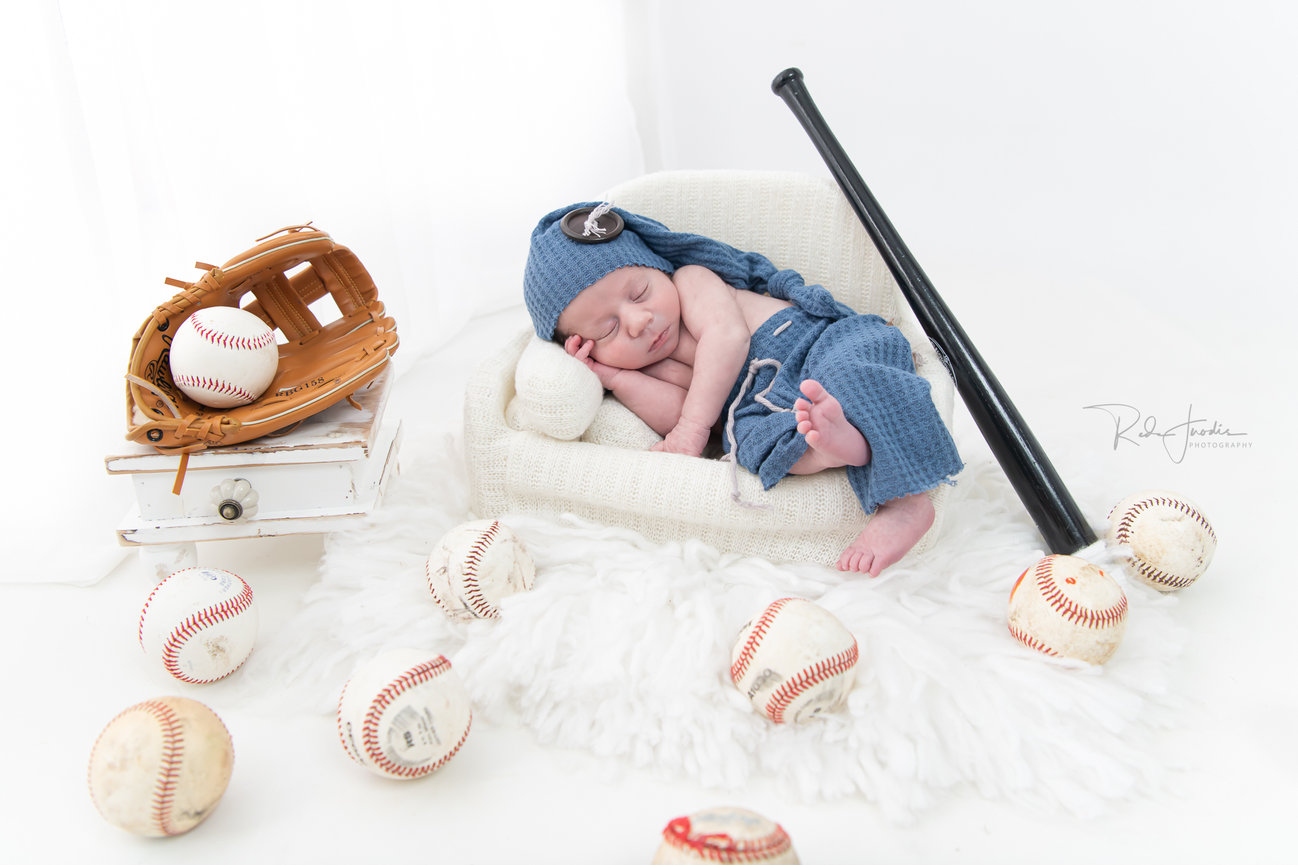 Newborn baby dressed in baseball attire sleeping on a soft surface, surrounded by baseballs and a glove.