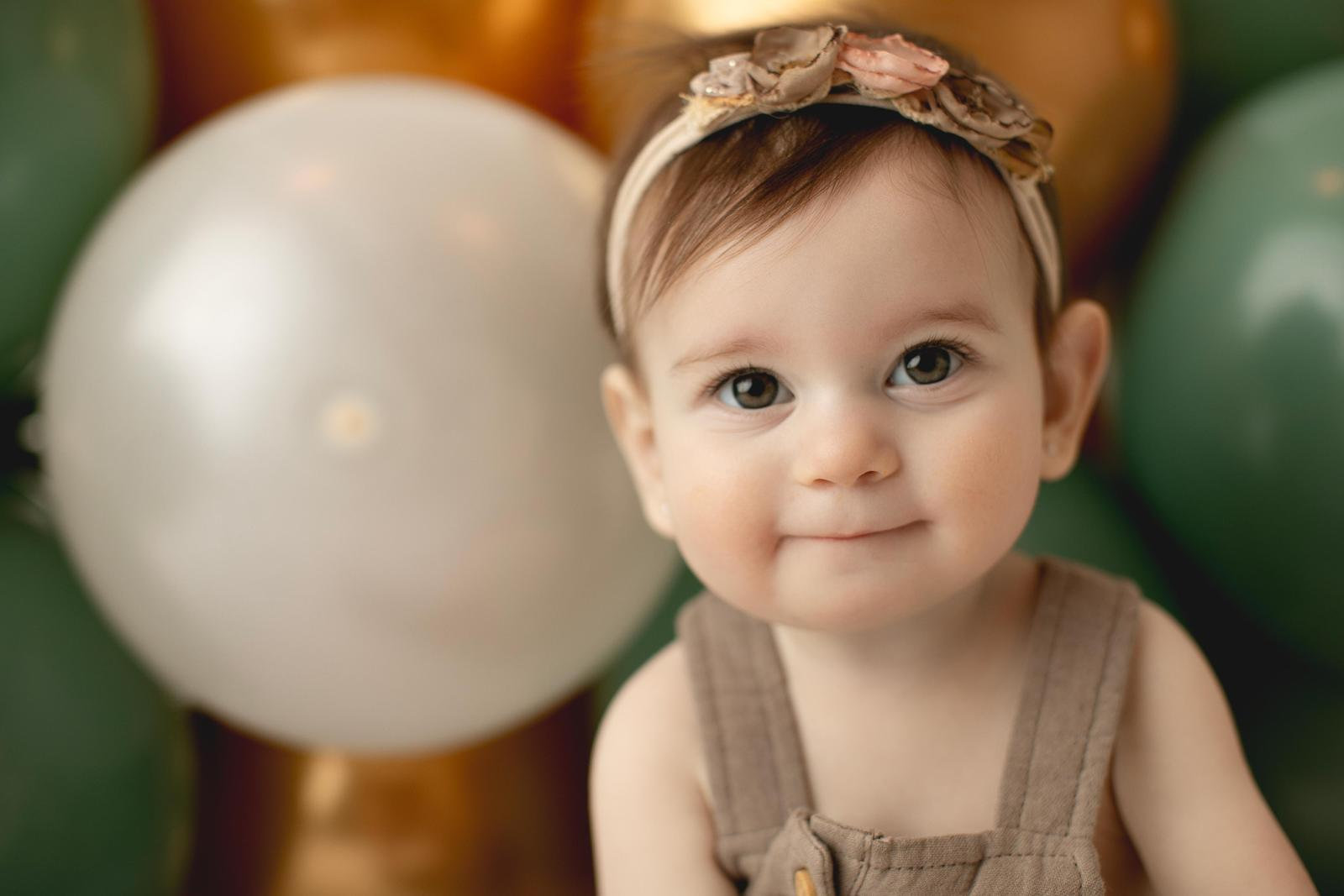 Baby sitting beside sage and gold balloons in a brown romper