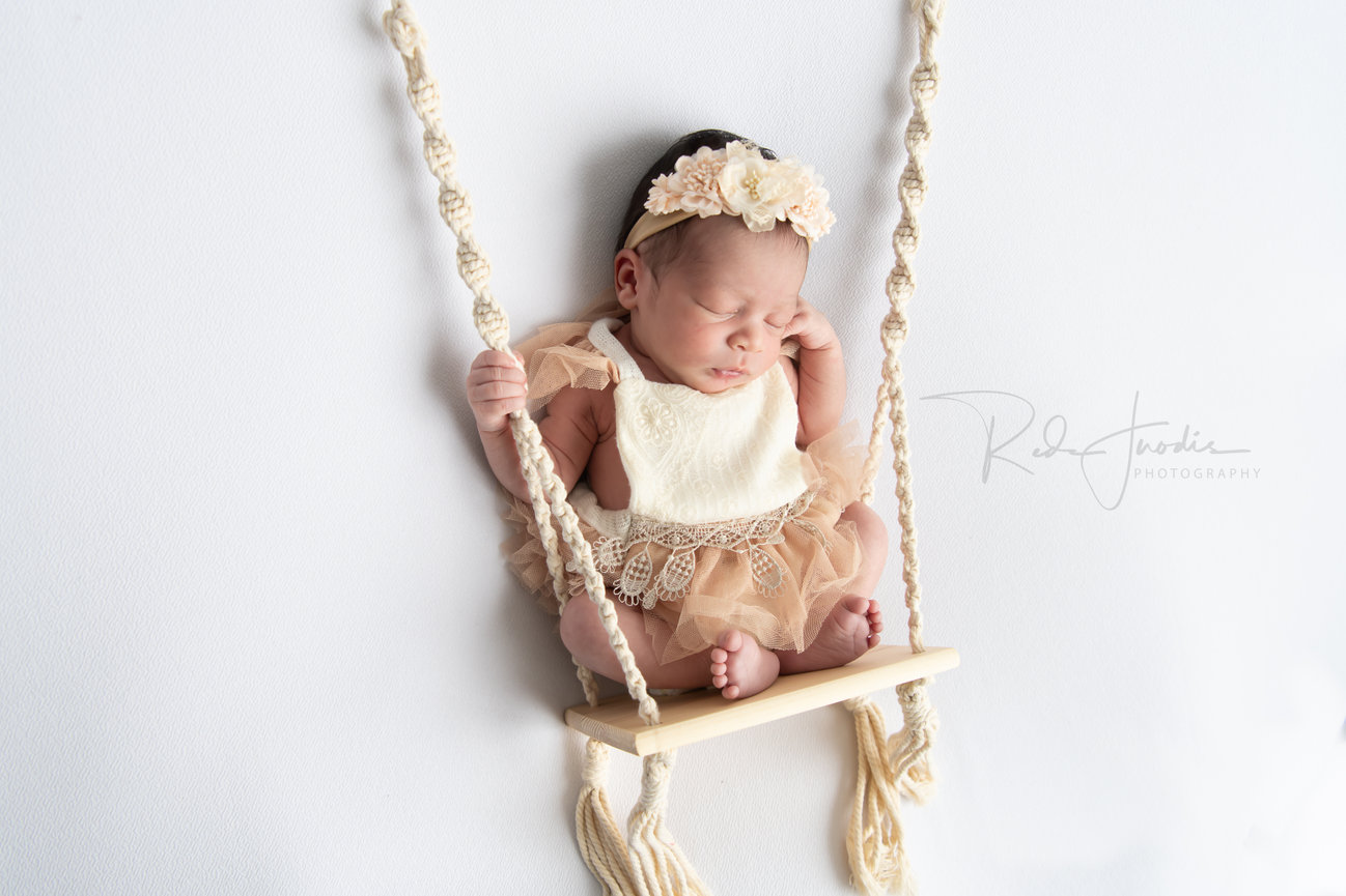 Newborn baby girl resting peacefully on a swing, wearing a floral headband and delicate outfit.