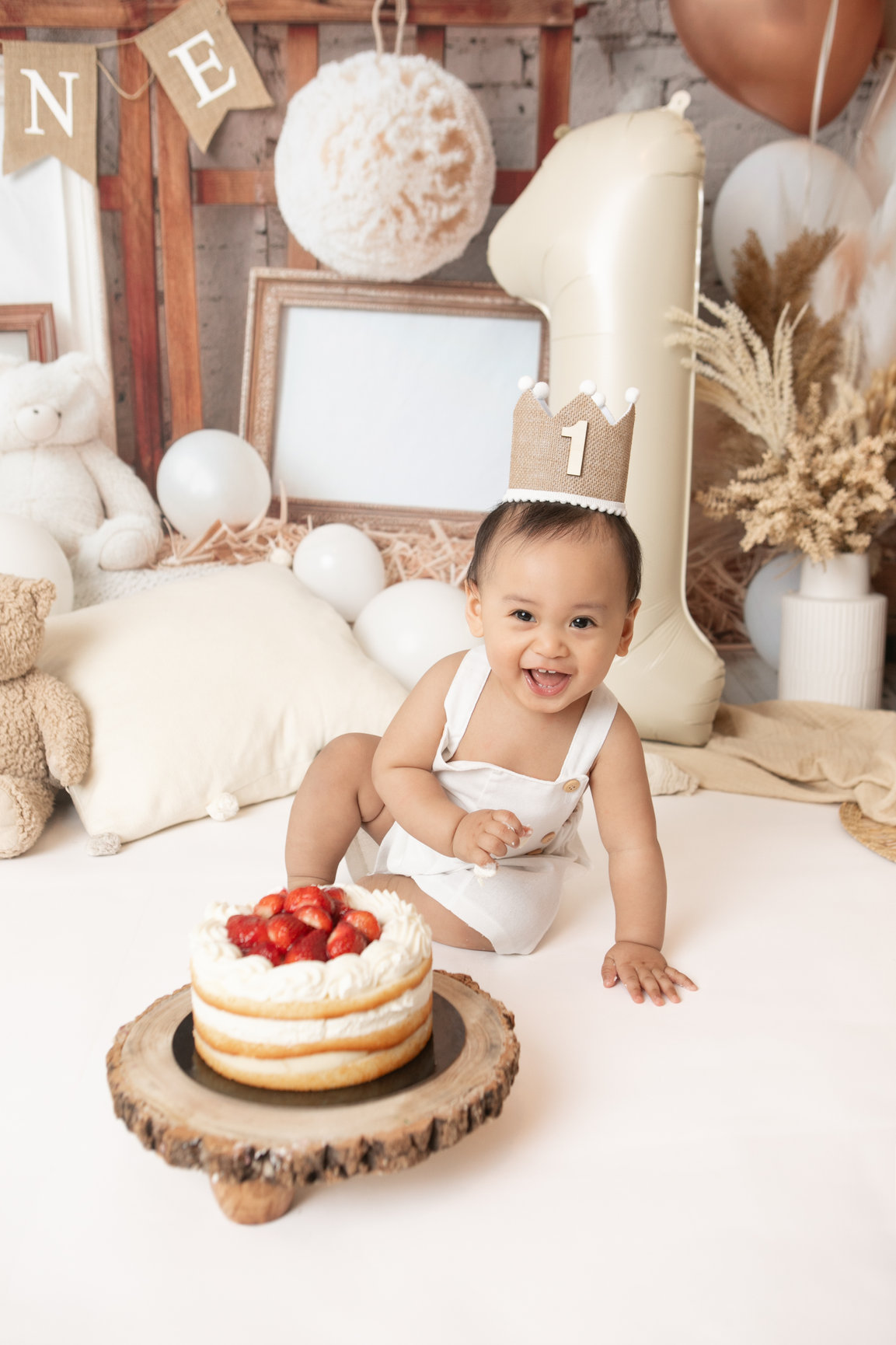 Smiling baby wearing a first birthday crown beside a strawberry-topped cake in a cream and ivory studio set