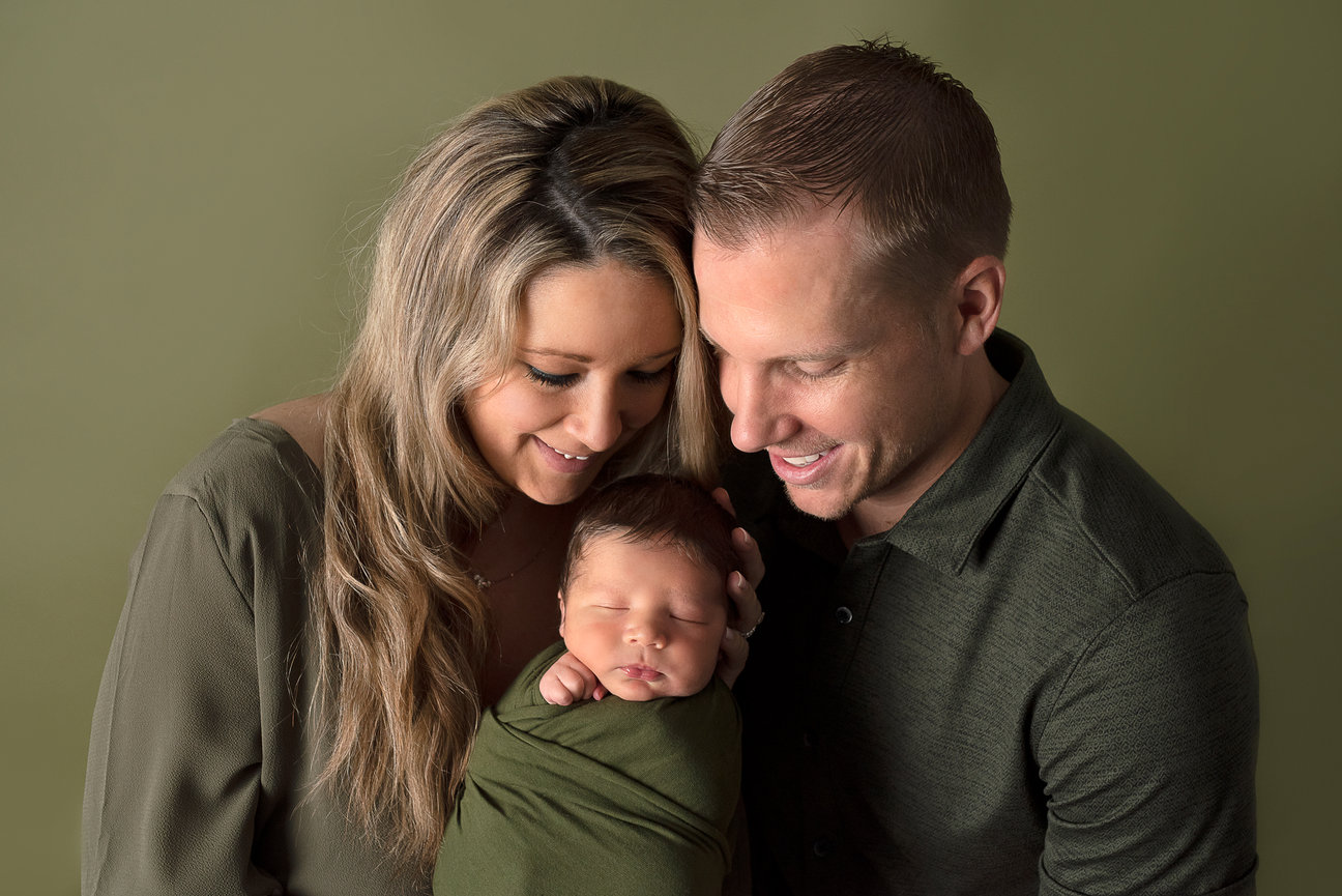 A couple smiles lovingly at a sleeping baby wrapped in a green blanket. All three are dressed in matching green tones, set against a plain olive background. The woman has long hair, both radiating happiness, in the Village of Lakewood, Illinois.