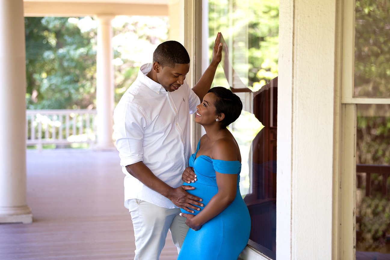 A couple stands on a porch, smiling at each other with unmistakable joy. The woman, in a blue dress, holds her pregnant belly as the man gently touches it. This serene moment of pregnancy is beautifully framed by the lush greenery in the background.