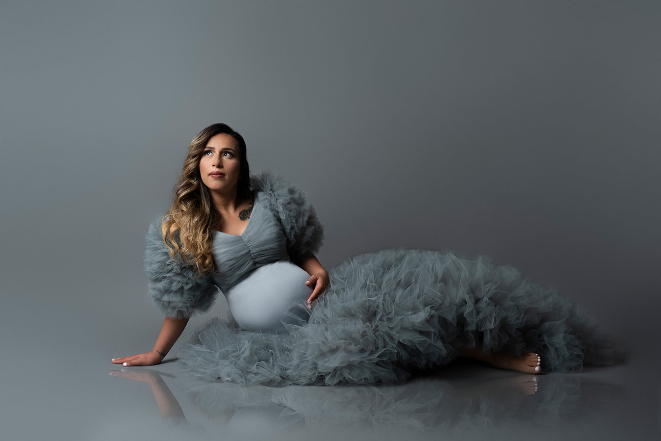 A pregnant woman is reclining on the floor, dressed in a flowing gray gown with ruffled sleeves. She gazes upward against a plain gray background. Her long hair cascades over her shoulder, and one hand rests gently on her belly.