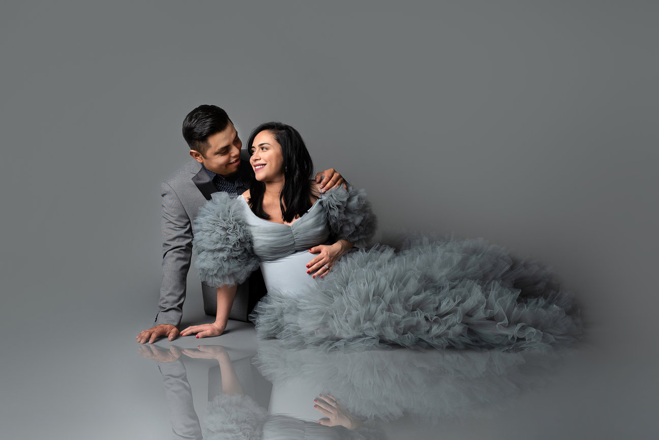A couple is smiling at each other, dressed in formal attire reminiscent of a baby and maternity photography session. The man is in a gray suit, while the woman wears a voluminous, feathered gray gown. They are on the floor against a gray backdrop.